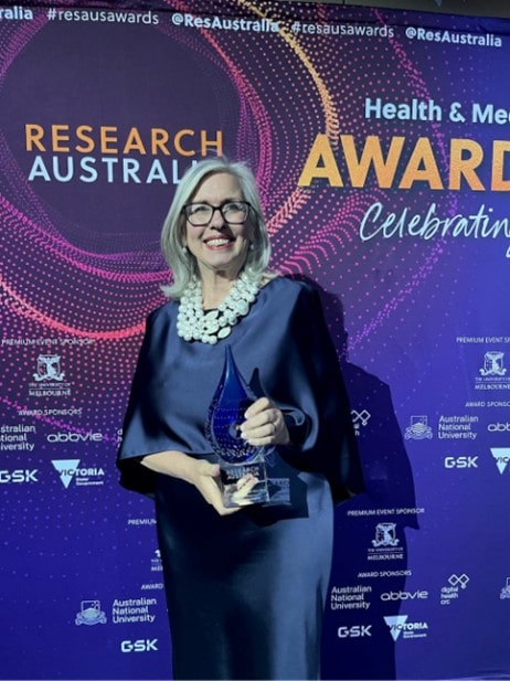 Professor Christobel Saunders AO holds her Health Services Research Award as she stands in front of a large purple banner reading ‘Research Australia’ at the 2024 Research Australia Health & Medical Research Awards 2024
