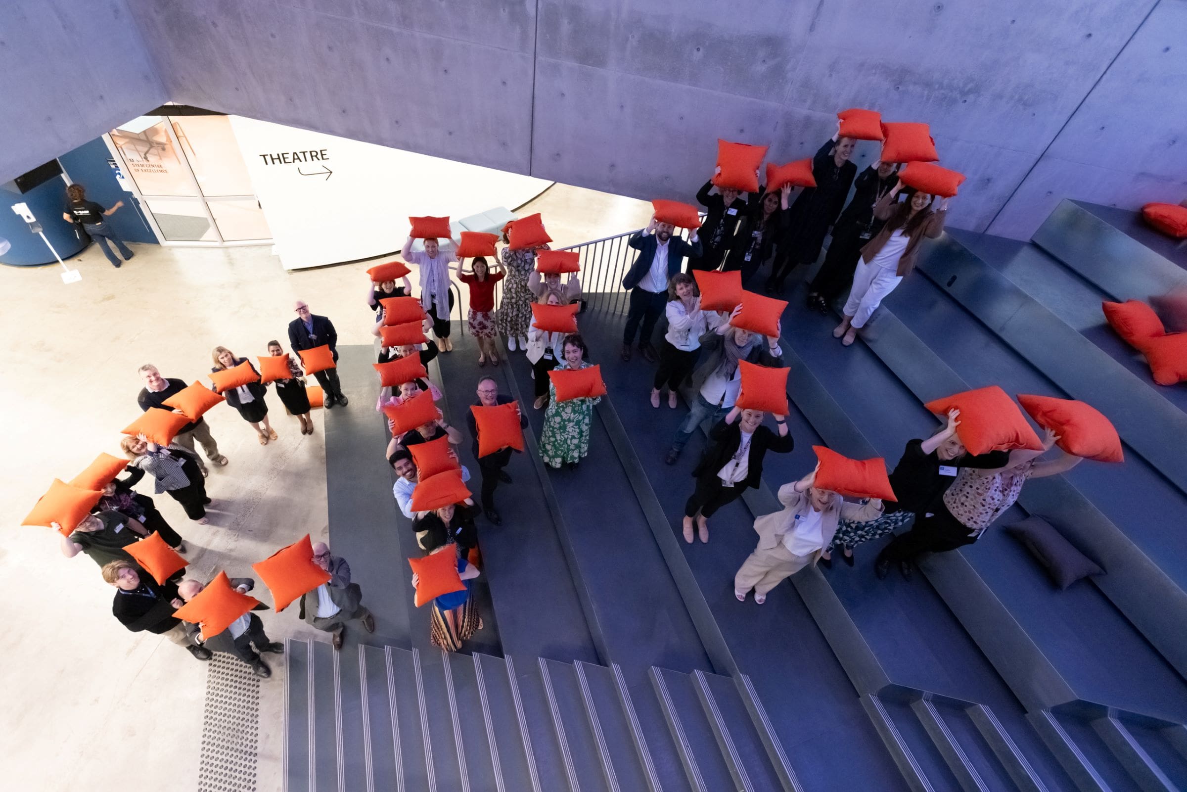 Faculty attendees at the launch event of the Collaborative Practice Centre assemble the letters ‘CPC’ holding orange cushions above their heads, on the steps inside the Science Gallery Melbourne