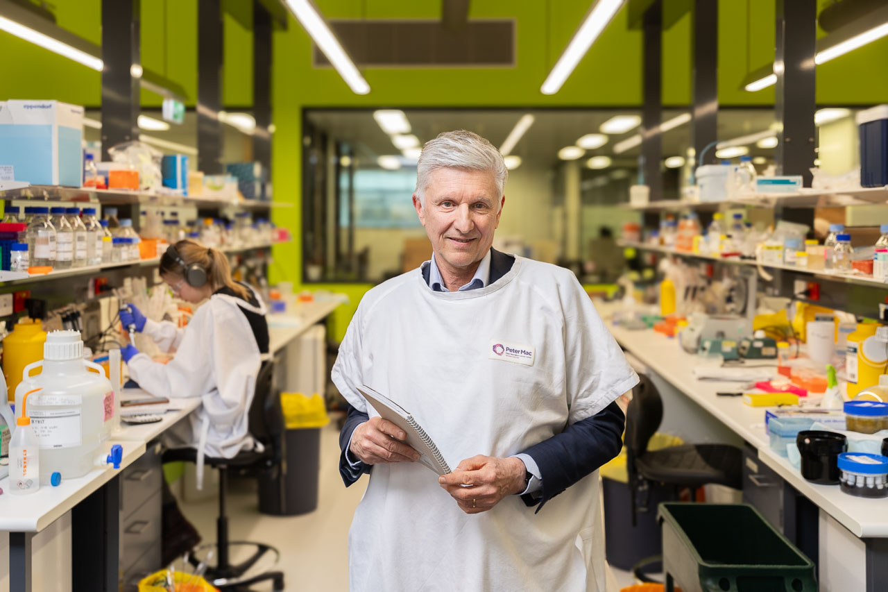 Alum Professor Grant McArthur AO stands in the middle of a research laboratory at the VCCC. A PhD student sits at a desk behind him wearing PPE and using a pipette