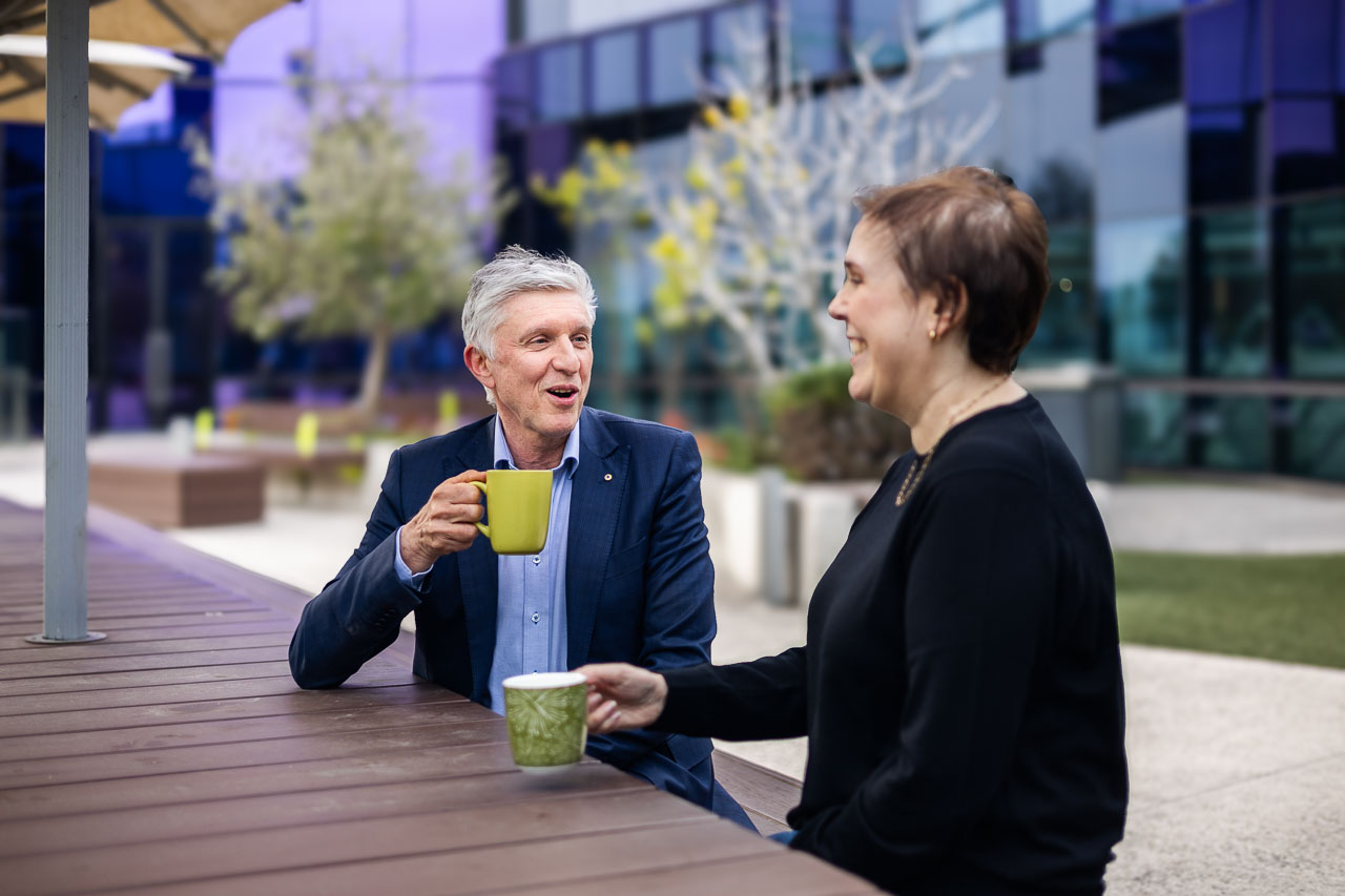 Alum Professor Grant McArthur AO, left, and previous patient and friend Melissa Sheldon, right, talk while drinking from coffee mugs on a long wooden table at the rooftop garden at the VCCC