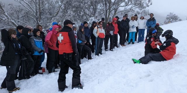 A row of medical students stand in the snow as the Falls Creek Ski Patrol demonstrate an on-slope rescue. Students are dressed for the cold, with fresh snow on the ground and overcast weather. 