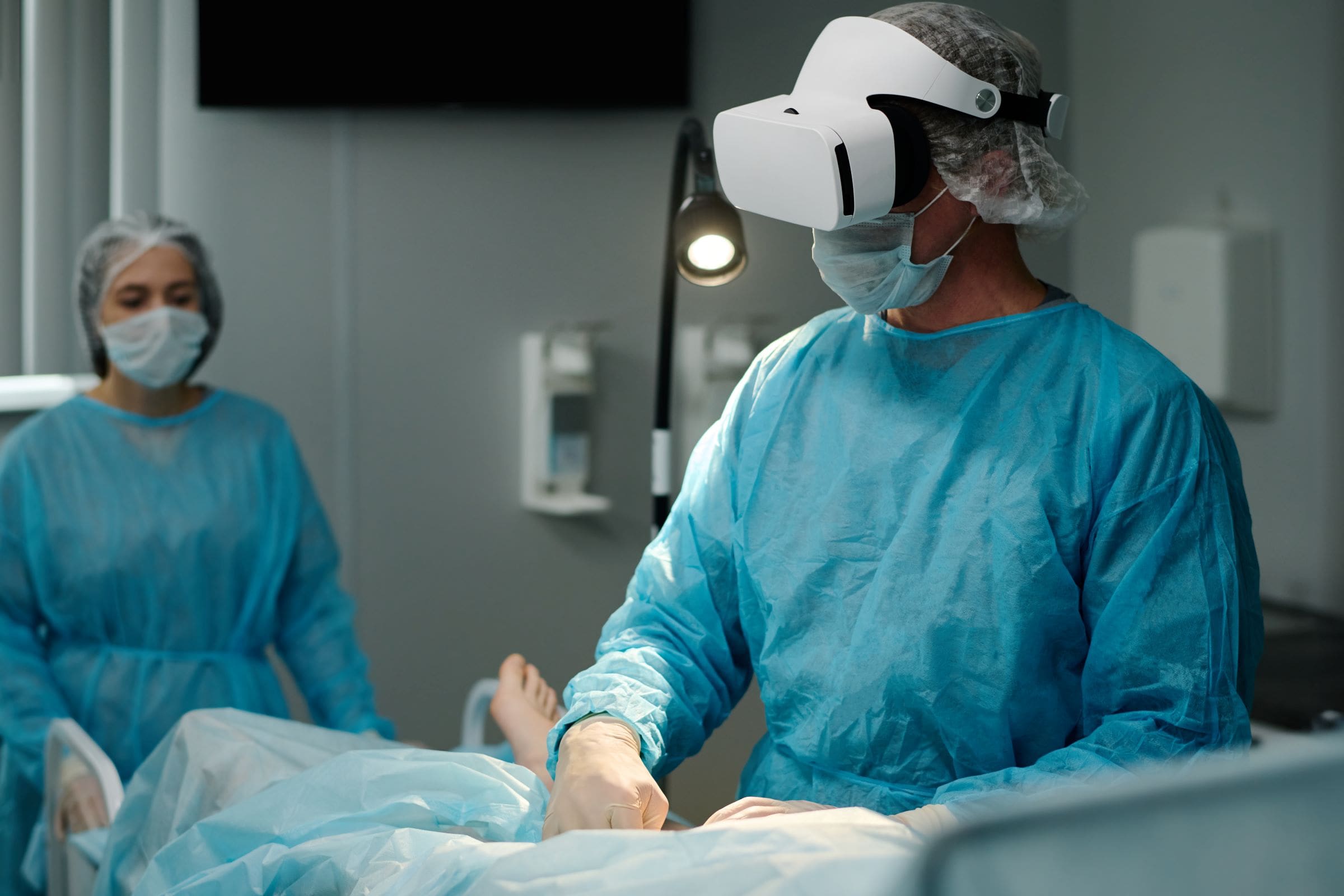 A surgeon wearing a surgical gown, mask, and Virtual Reality headset performs a simulated procedure, while another clinician in scrubs and mask observes in the background