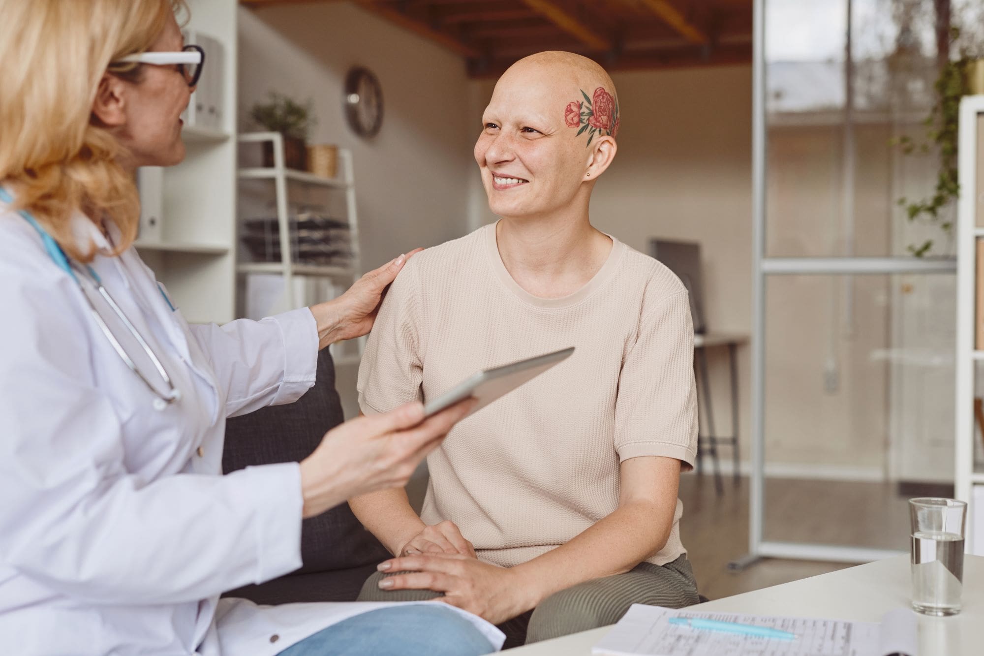 A clinician holding a tablet, left, puts her hand on the shoulder of a bald cancer patient during a clinic visit. They smile at each other.