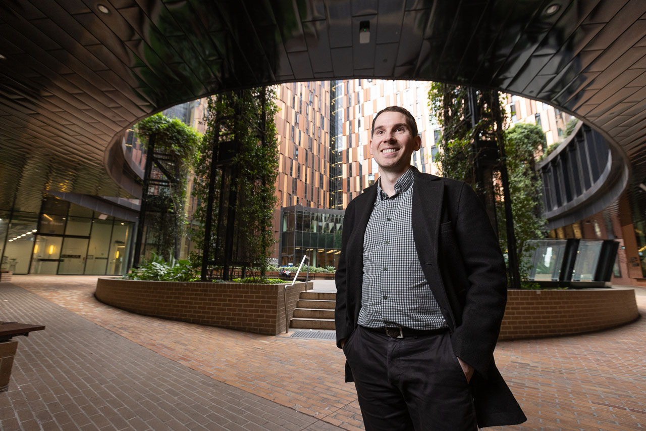Alum Dr Emerson Keenan stands smiling at the Womin-Djerring, a brightly lit open-air oculus with draping green vines at the heart of Melbourne Connect